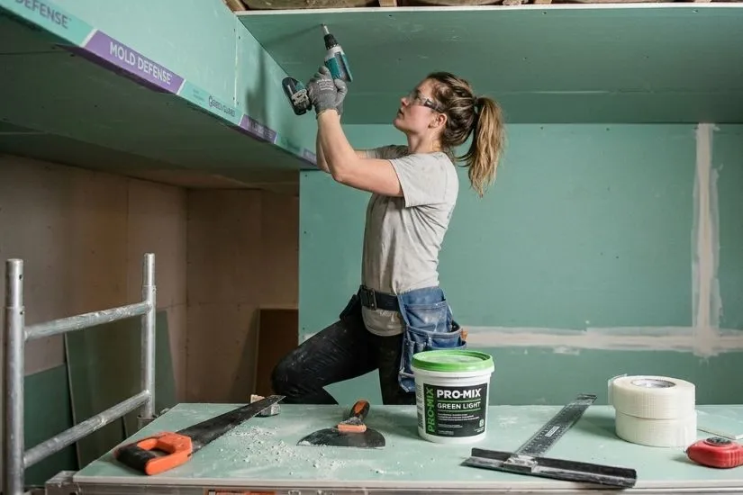 A professional contractor installing moisture-resistant green board drywall on a basement wall using a power drill.